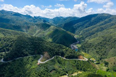 Laos 'taki Phou Khoun ve Luang Prabang arasında yoğun yeşil tepeler ve vadilerden geçen eğimli bir dağ yolunun hava manzarası. Manzarada katmanlı tepeler, dağınık kırsal binalar ve dramatik bulut gölgeleri yer alıyor.