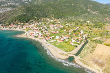 Kato Vasiliki 'nin yüksek açılı görüntüsünde, turkuaz sular ve parlak güneş ışığı altında yemyeşil tepelerle çevrili kıvrımlı bir kıyı şeridi boyunca uzanan kırmızı çatılı evler görülüyor. Manzara küçük bir kanal, açık alanlar ve sakin bir kanala sahiptir.
