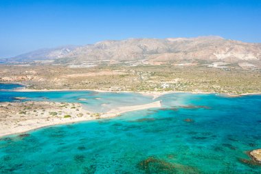 Yunanistan 'ın Girit kentindeki Elafonisi plajının havadan görünüşü, turkuaz sığ ve engebeli, güneşli tepeler arasında uzanan çarpıcı bir kum çubuğuyla. Sakin sahnede canlı mavi su, yumuşak kumlu dokular ve açık Akdeniz manzarası yer alıyor..