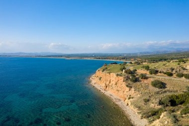 Turuncu kayalıkların şeffaf mavi suyla buluştuğu engebeli bir sahil manzarası. Açık kırsal ve bulutlu bir gökyüzünün altındaki uzak dağlarla çevrili. Güneş ışığı Akdeniz 'in canlı renklerini ve doğal dokularını güçlendirir