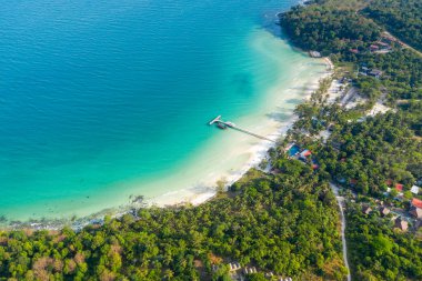 Tepedeki perspektif, Kamboçya 'nın Koh Rong adasındaki Coconut Sahili' nde beyaz kumdan canlı turkuaz denize uzanan uzun bir iskele olduğunu gösteriyor. Yoğun tropikal ormanlar, dağınık binalar ve nazik dalgalar hareketli ve sakin bir tatil beldesi oluşturur.