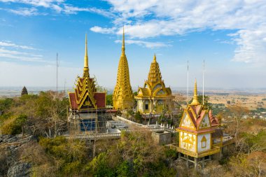 Altın pagodalar ve tapınak yapıları Phnom Sampov 'un kayalık zirvesinin üzerinde yükselirler, Kamboçya' nın Battambang yakınlarındaki geniş kırsal düzlüklere karşı kurulurlar. Sahne Khmer mimarisi, kurak mevsim yeşillikleri ve geniş bir alanı vurguluyor.