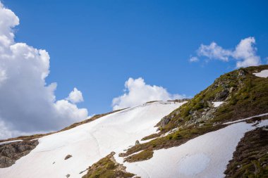 Aiguillette des Houches 'ta güneşli bir dağda eriyen karlar, açıktaki kayalar ve dağınık kümülüs bulutları ile canlı mavi gökyüzünün altında seyrek alp bitkileri sergilenir..