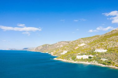 Bereketli yeşil tepelerin, canlı bir gökyüzünün altında berrak mavi denizle buluştuğu kıyı şeridi boyunca uzanan dağınık beyaz binaların olduğu parlak kıyı manzarası. Atmosfer taze, açık ve davetkar. Yumuşak güneş ışığı doğal dokuları aydınlatıyor..