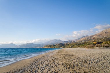 Geniş kumlu kıyı şeridi turkuvaz suları boyunca uzanır ve parlak, berrak bir gökyüzünün altında engebeli dağlar yükselir. Huzurlu kıyı manzarası, huzurlu ve açık bir atmosferi çağrıştıran sıcak güneş ışığıyla yıkanır..
