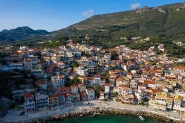 Yoğun ormanlık tepeler ve engebeli dağlara karşı Yunanistan 'ın Parga kentinin rıhtımında kırmızı kiremitli çatıları olan renkli evler. Parlak gün ışığı ve açık gökyüzü Akdeniz mimarisi ve canlı kıyı manzarasını vurguluyor.