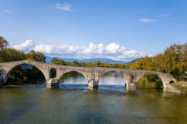 Yunanistan 'ın Arta kentinde açık bir nehir boyunca uzanan tarihi çok kemerli taş köprünün panoramik manzarası. Sonbahar ağaçları, uzak dağlar ve bir kemerin altındaki Yunan bayrağı mavi gökyüzünün altındaki bu sakin sahneyi tamamlıyor..