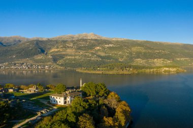 Altın sabah ışığı, arka planda Pindus dağlarının orman yamaçlarının yükseldiği Ioannina Gölü kıyısında bir göl camii ve tarihi bir binayı ön plana çıkarıyor. Sakin su ve berrak mavi gökyüzü sakin bir doğal ortam yaratır.