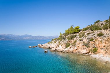 Yeşil çalıları ve çam ağaçları olan güneşli kayalık kıyı şeridi bulutsuz bir gökyüzünün altında şeffaf turkuaz denizle buluşuyor. Uzak dağlar Yunanistan 'ın Nafplio kentinde sakin bir Akdeniz zemin yarattı.