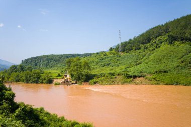 Sarı bir kazıcı, yemyeşil tepeler ve yoğun bitki örtüsüyle çevrili çamurlu nehir kıyısı boyunca çalışıyor. Kuzey Vietnam 'da parlak mavi bir gökyüzü altında. Güneş ışığı kırsal araziyi aydınlatır, yeryüzü ile canlı arasındaki zıtlığı vurgular.