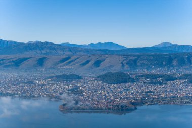 Geniş panoramik manzara, Yunanistan 'ın Epirus kentindeki Pindus dağlarının engebeli yamaçlarına karşı kurulmuş Ioannina kentini ve Ioannina Gölü' ndeki adasını yakalıyor. Sis zerreleri durgun suyun üzerinde sürükleniyor.