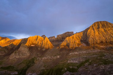 Sıcak güneş ışığı, La Breche de Roland 'ın engebeli kaya oluşumlarını yukarıdaki dramatik gökyüzü ile çelişen altın bir parıltıyla yıkar. Sahne, Fransız Pireneleri 'ndeki dokulu katman ve kalın dağ şekillerini vurguluyor..