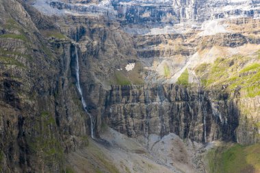 İnce bir şelale, Cirque de Gavarnie 'deki yüksek, yıpranmış kaya duvarlarından aşağıya katmanlı taş dokularla ve yumuşak gün ışığı altında seyrek yeşillikle düşer..