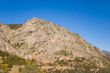 Orta Yunanistan 'da canlı bir mavi gökyüzüne karşı kurulmuş, çıplak kaya yüzleri ve dağınık yeşil bitki örtüsüne sahip, güneşli, engebeli bir dağ. Manzara, dokulu uçurumlar, seyrek ağaçlar ve sakin bir doğal atmosfere sahiptir..