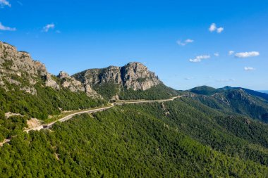 Sardinya 'nın Baunei ve Dorgali yakınlarındaki yemyeşil tepeler ve kayalık uçurumlar boyunca uzanan dolambaçlı bir yol. Parlak güneş ışığı, parlak mavi gökyüzünün altındaki sık ormanı ve engebeli dağ dokusunu vurgular..