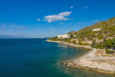 Parlak mavi bir gökyüzünün altında yeşil tepelerle çevrili kayalık bir kıyı şeridi ve turkuaz deniz üzerinde tünemiş sakin bir sahil manzarası. Güneş ışığı manzaranın dokusunu güçlendirir, barışçıl ve davetkar bir atmosfer yaratır..