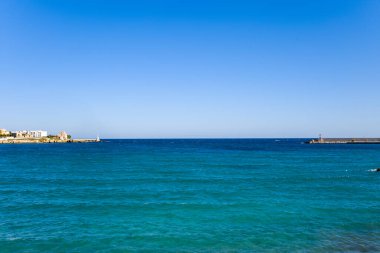 Canlı turkuaz sular Adriyatik Denizi boyunca uzanır, Otrantos limanı ve bulutsuz mavi gökyüzünün altında görünen uzak deniz feneri ile. Sahne, İtalya 'nın güneyindeki berrak kıyı suyu, kentsel kıyı şeridi ve açık ufuk arasındaki zıtlığı vurguluyor.