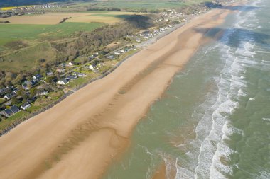 Hava görüntüsünde, Omaha Sahili 'ndeki Normandiya kıyısı boyunca hafif dalgalarla çevrili geniş bir altın kum tabakası görülüyor. Sahne güneşli, yeşil alanlar ve huzurlu bir kıyı şeridi var..