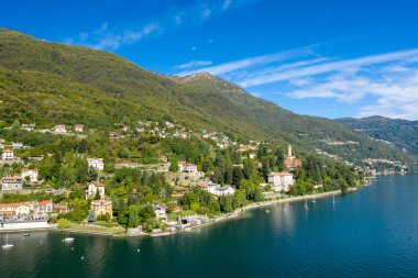 Kuzey İtalya 'daki Maggiore Gölü' nün yeşil tepeleri boyunca uzanan pitoresk bir göl köyünün panoramik manzarası. Sahnede geleneksel evler, yemyeşil bitki örtüsü ve berrak bir gökyüzü altında sakin mavi su, sakin bir alp manzarası yaratıyor..