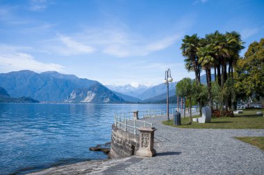 İtalya 'nın Verbano kentindeki rıhtım boyunca palmiye ağaçları ve klasik sokak lambalarıyla kaplı eğimli taş kaldırım. Manzara, Maggiore Gölü 'nün sakin mavi sularına, berrak bir gökyüzünün altındaki uzak dağlara, Akdeniz ve Alp elementlerinin karışımına bakıyor.