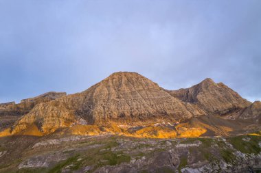 Sıcak akşam ışığı, Pireneler 'deki engebeli bir dağ yüzeyinin dokulu kaya katmanlarını aydınlatır. Yumuşak, bulutlu bir gökyüzü altında dramatik manzarada altın renkler bırakır..