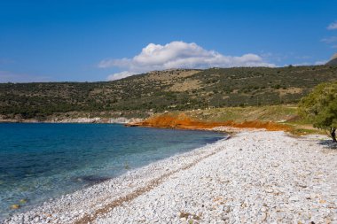 Turkuaz suyla karşılaşan beyaz çakıl taşı kıyı şeridi geniş bir görüş alanına sahip. Turuncu kayalıklar ve parlak mavi gökyüzünün altındaki yeşil tepeler. Güneş ışığı, doğal dokuları ve canlı kıyı renklerini vurgular.