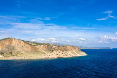 Monemvasias 'ın engebeli kayalıkları ve canlı mavi Akdeniz sularıyla çevrili antik taş yerleşim yerlerini geniş bir gökyüzü altında dağınık bulutlarla birlikte. Dramatik manzarada desenli kaya yüzeyleri, kıyı yeşilliği ve geniş genişlik yer alıyor.