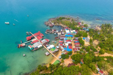 Hava görüntüsü, Coconut Beach, Koh Rong, Kamboçya 'da canlı çatıları ve turkuaz suya uzanan ahşap iskelesi olan renkli bir balıkçı köyü gösteriyor. Palmiye ağaçları, yemyeşil ve kayalık kıyı şeridi Bu sakin kıyı manzarası.