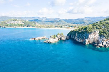 Yunanistan 'ın Epirus kentindeki Karavostasi kıyı şeridi boyunca uzanan engebeli kayalık kayalıkları ve çıkıntıları çevreleyen canlı mavi sular görülüyor. Parlak gün ışığı bu geniş Akdeniz deniz manzarasında yemyeşil tepeler ve kumlu bir plajı gözler önüne seriyor..
