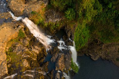 Hava perspektifi, Laos 'un güneyindeki Tad Yuang şelalesindeki engebeli bazalt kayaların üzerinden akan suyu yakalar. Sıcak güneş ışığı, orman yamaçlarındaki dokulu taşları ve yemyeşil yeşillikleri vurguluyor..
