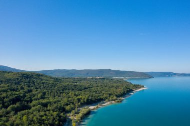 Lac de Sainte-Croix 'in hava manzarası yoğun yeşil ormanlarla çevrili ve bulutsuz mavi gökyüzünün altında yuvarlanan tepelerle çevrili geniş bir turkuaz kıyı şeridini ortaya çıkarıyor..