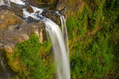 Yakın çekim havadan bakıldığında Tad Yuang şelalesinin engebeli bazalt kayaların üzerinden Güney Laos 'taki Bolaven Platosu' nda yoğun yeşil yapraklara daldığını görüyoruz. Ilık güneş ışığı, taşın, akan suyun ve canlı bitki örtüsünün zıt dokularını güçlendirir..
