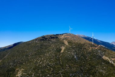 Yunanistan 'ın Thisbe kenti yakınlarındaki açık mavi bir gökyüzünün altındaki engebeli, yeşil bir yamaçta üç rüzgar türbini duruyor. Manzara seyrek bitki örtüsü, toprak dokular ve parlak gün ışığında dolambaçlı toprak yollar içerir..