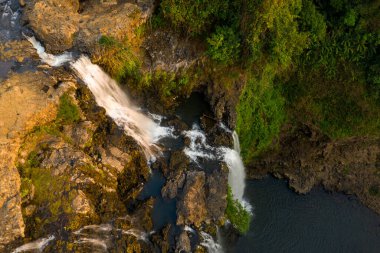 Yukarıdan bakıldığında, Tad Yuang şelaleleri engebeli bazalt kayalarının üzerinden, Laos 'un güneyindeki Bolaven Platosu' ndaki yemyeşil bitki örtüsüyle çevrili karanlık bir havuza dönüşür. Sıcak güneş ışığı kayalık arazinin dokusunu güçlendirir ve kayanın hareketini vurgular.