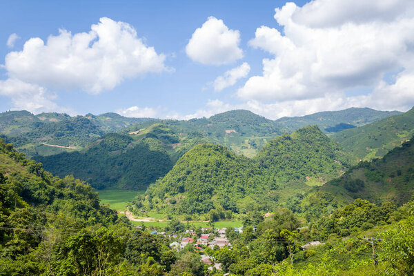 A panoramic view of vibrant green hills and a small rural village nestled in a sunlit valley under a bright blue sky with scattered clouds in northern Vietnam. The landscape is lush, expansive, and peaceful, with dense foliage and rolling mountains.