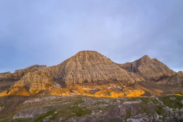 Sıcak akşam ışığı, Pireneler 'deki engebeli bir dağ yüzeyinin dokulu kaya katmanlarını aydınlatır. Yumuşak, bulutlu bir gökyüzü altında dramatik manzarada altın renkler bırakır..