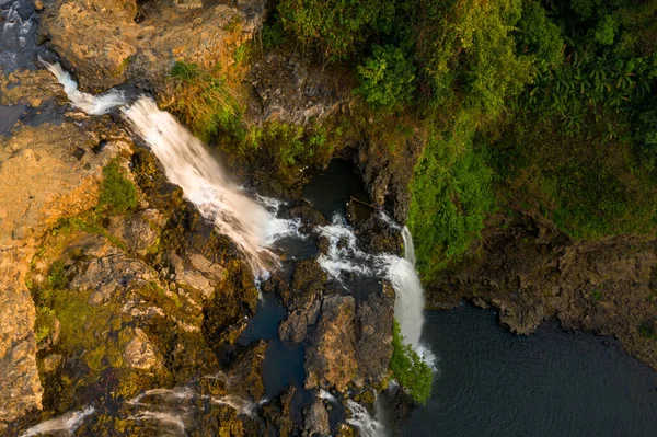 Yukarıdan bakıldığında, Tad Yuang şelaleleri engebeli bazalt kayalarının üzerinden, Laos 'un güneyindeki Bolaven Platosu' ndaki yemyeşil bitki örtüsüyle çevrili karanlık bir havuza dönüşür. Sıcak güneş ışığı kayalık arazinin dokusunu güçlendirir ve kayanın hareketini vurgular.