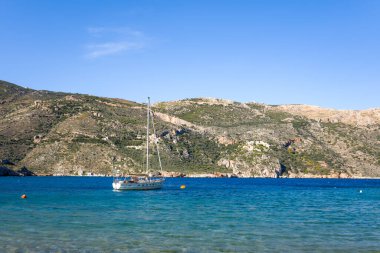 Yunanistan 'ın Porto Kagio kentinin engebeli tepeleri yakınlarında, kayalık kıyı şeridini aydınlatan açık mavi bir gökyüzünün altında, beyaz bir yelkenli turkuaz su üzerinde yüzüyor..