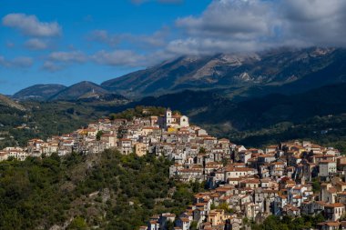 İtalya 'nın Rivello kenti yoğun evleri ve dramatik dağlarla çevrili merkezi bir kilisesi olan yemyeşil bir yamacın tepesinde, gün ışığı altında dağılmış bulutlarla oturmaktadır..