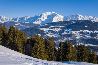 Karla kaplı yamaçlar, Megeve yakınlarında açık mavi bir gökyüzünün altında yükselen görkemli Mont Blanc Massif ile yoğun bir çam ormanına doğru iniyor..