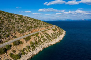 Peloponnese kıyı şeridine bakan, seyrek bitki örtüsüyle kaplı kayalık bir yamaç boyunca uzanan kıvrımlı bir yol beyaz bulutlarla kaplı bir gökyüzünün altında. Sahne özgürlük ve manzara yolculuğunu çağrıştırıyor..