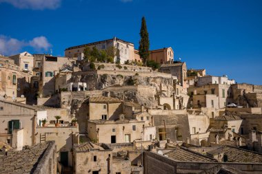 Kümelenmiş taş evler Matera, İtalya 'da kayalık bir yamaç boyunca yükseliyor, dokulu cepheleriyle ve parlak mavi gökyüzünün altında selvi ağacıyla.