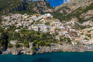 Renkli Akdeniz binaları Positano, İtalya 'nın dik yamaçlarından aşağı akarak turkuaz sulara ve engebeli uçurumlara bakıyor. Bereketli yeşillik ve tarihi bir taş gözetleme kulesi açık mavi gökyüzünün altındaki dramatik kıyı manzarasını ön plana çıkarıyor..