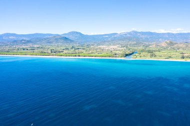 Lotzorai, Sardinya 'nın yüksek açılı manzarası, canlı turkuaz Akdeniz suları, dar kumlu bir kıyı şeridi ve berrak mavi gökyüzünün altında uzak dağları olan yeşil alanlar gösteriyor. Sahne parlak, açık ve sakin..