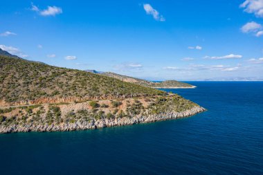 A rugged peninsula with rocky slopes and sparse vegetation juts into the deep blue waters under a bright sky. The vivid contrast between land and sea creates a sense of tranquility and natural beauty.