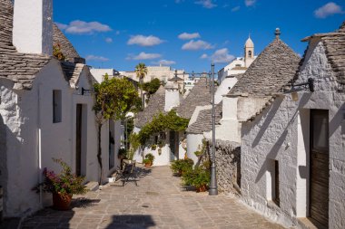 İtalya 'da Alberobello, Apulia' da beyazlatılmış trullo evleri ve taş çatılarla dolu büyüleyici dar bir cadde. Saksı bitkileri, kırsal dokular ve berrak mavi gökyüzü canlı bir güney İtalya atmosferi yaratır..