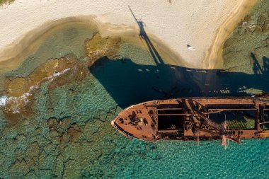 Dimitrios 'un enkazının tepeden görünüşü, Yunanistan' ın Gythio kenti yakınlarındaki altın kum ve sığ turkuaz suya dramatik bir gölge düşürüyor. Güneş ışığı paslı metal dokuları, kayalık deniz yatağını ve gemi, deniz ve plaj arasındaki canlı kontrastı ortaya çıkarır..