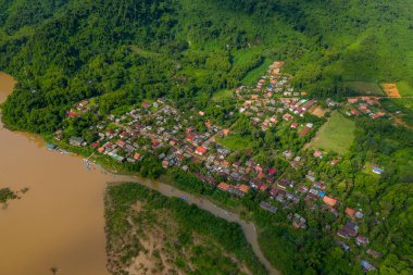 Muang Ngoi köyünün yüksek açılı panoramik manzarası yemyeşil ormanla kuzey Laos 'taki çamurlu Nam Ou nehri arasında yer almaktadır. Görüntü renkli çatıları, nehir kenarındaki tekneleri ve sarp dağlarla çevrili canlı bitkileri ön plana çıkarıyor..