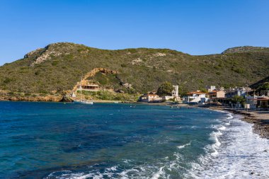 Dalgalar, Yunanistan 'ın Porto Kagio kentindeki küçük bir kıyı köyünün yanındaki çakıl taşlı bir kıyı şeridine vuruyor ve açık mavi gökyüzünün altında engebeli yeşil tepeler yükseliyor..