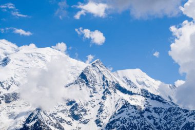 Aiguille de Bionnassay 'in keskin karla kaplı zirvesi engebeli dağlık yamaçların üzerinde kısmen bulutlarla kaplı parlak mavi gökyüzünün altında yükselir..